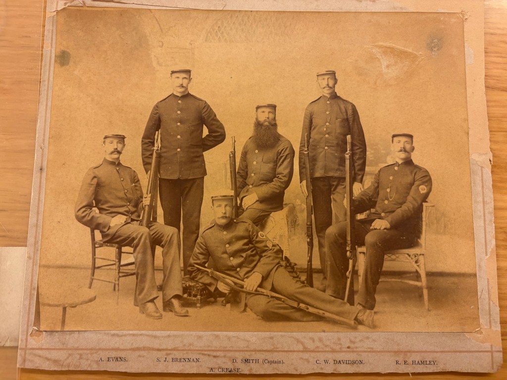 A historic photograph showing six men in military uniforms, posed in a studio setting. Five men stand while one is sitting with a rifle across his lap. The image has a vintage appearance, with a faded background.