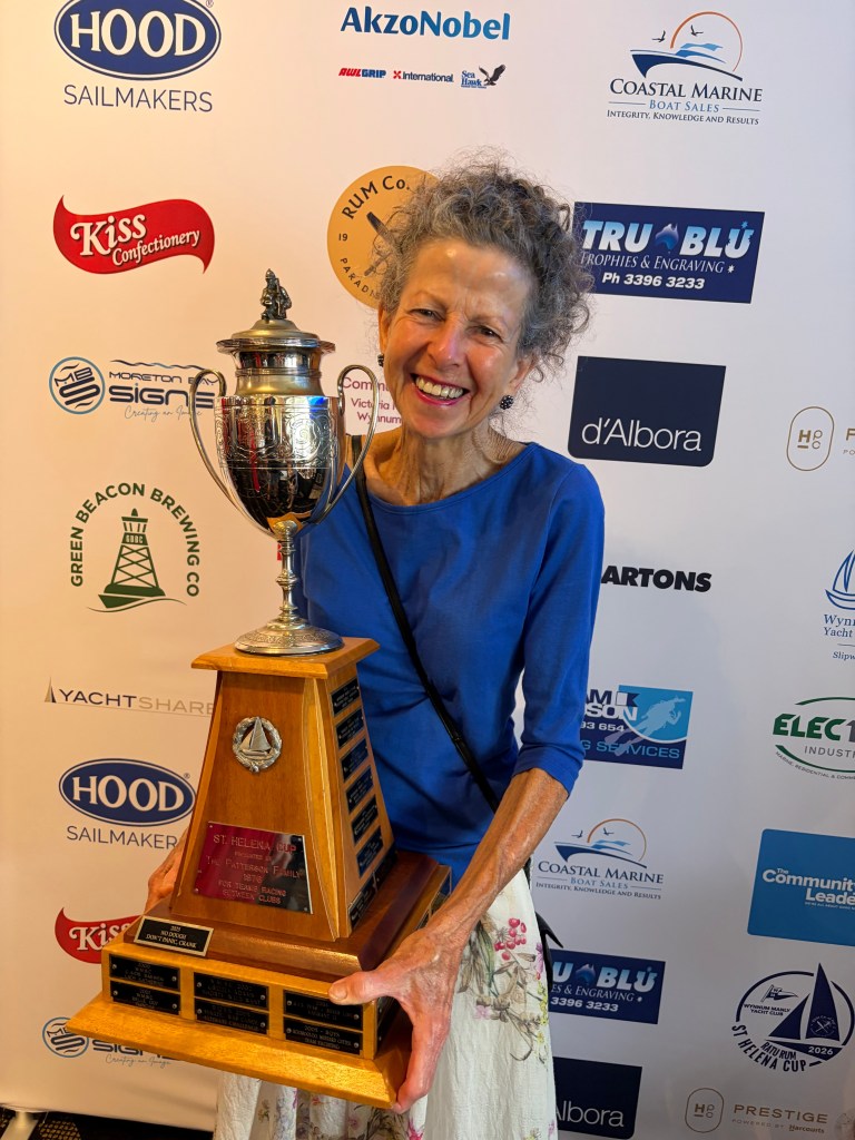 A smiling woman in a blue top holds a large trophy with a sailboat emblem, standing in front of a backdrop featuring various brand logos.