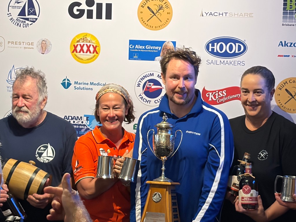 A group of four people celebrating with a trophy and drinks, standing in front of a branded backdrop featuring various sponsors of the St Helena Cup sailing event.