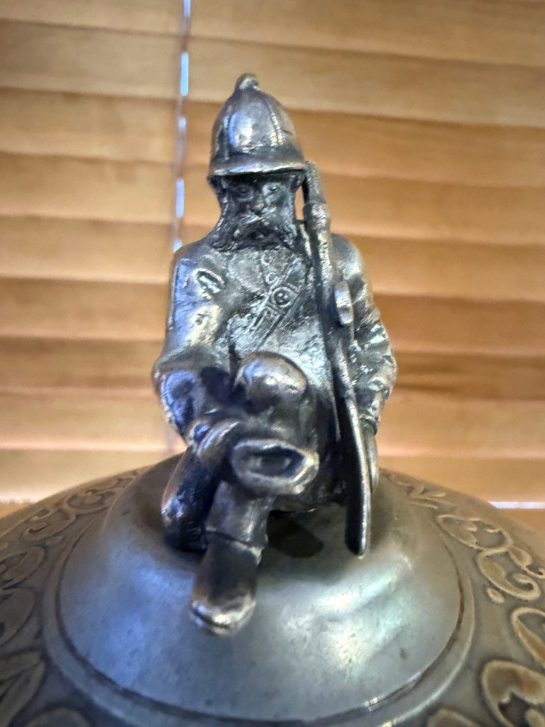 A close-up of a decorative metal figurine depicting a seated firefighter with a helmet and a hose, placed atop an ornate circular base.