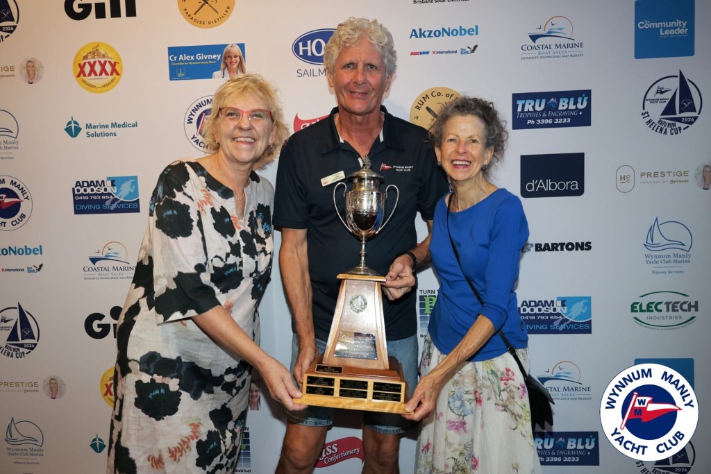 Three individuals holding a trophy at a yacht club event, with a backdrop featuring various sponsor logos.