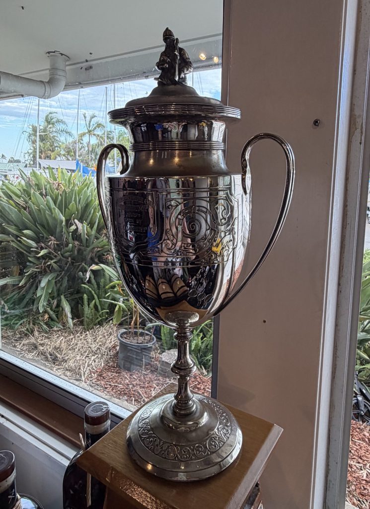 A detailed silver trophy on display, featuring ornate engravings and a small figure atop, set against a window with a view of green foliage and boats outside.